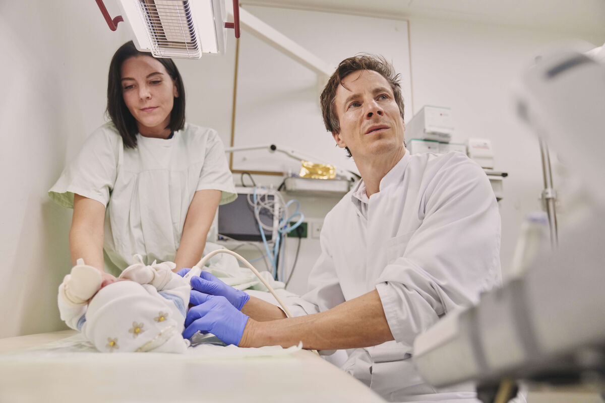 Das Bild zeigt einen Arzt und eine Mitarbeiterin bei der Untersuchung eines Neugeborenen.