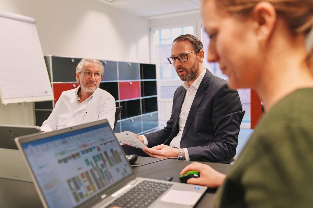 Das Bild zeigt einen Arzt, eine am Notebook arbeitende Mitarbeiterin und einen Mitarbeiter.