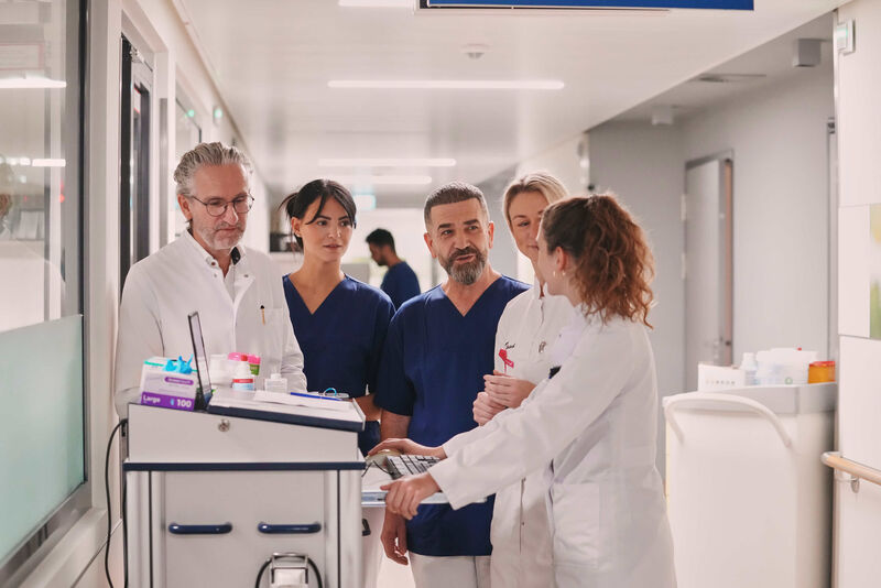 Das Bild zeigt einen Arzt und zwei Ärztinnen auf Station im Gespräch mit einer pflegerischen Mitarbeiterin und einer männlichen Pflegefachkraft.